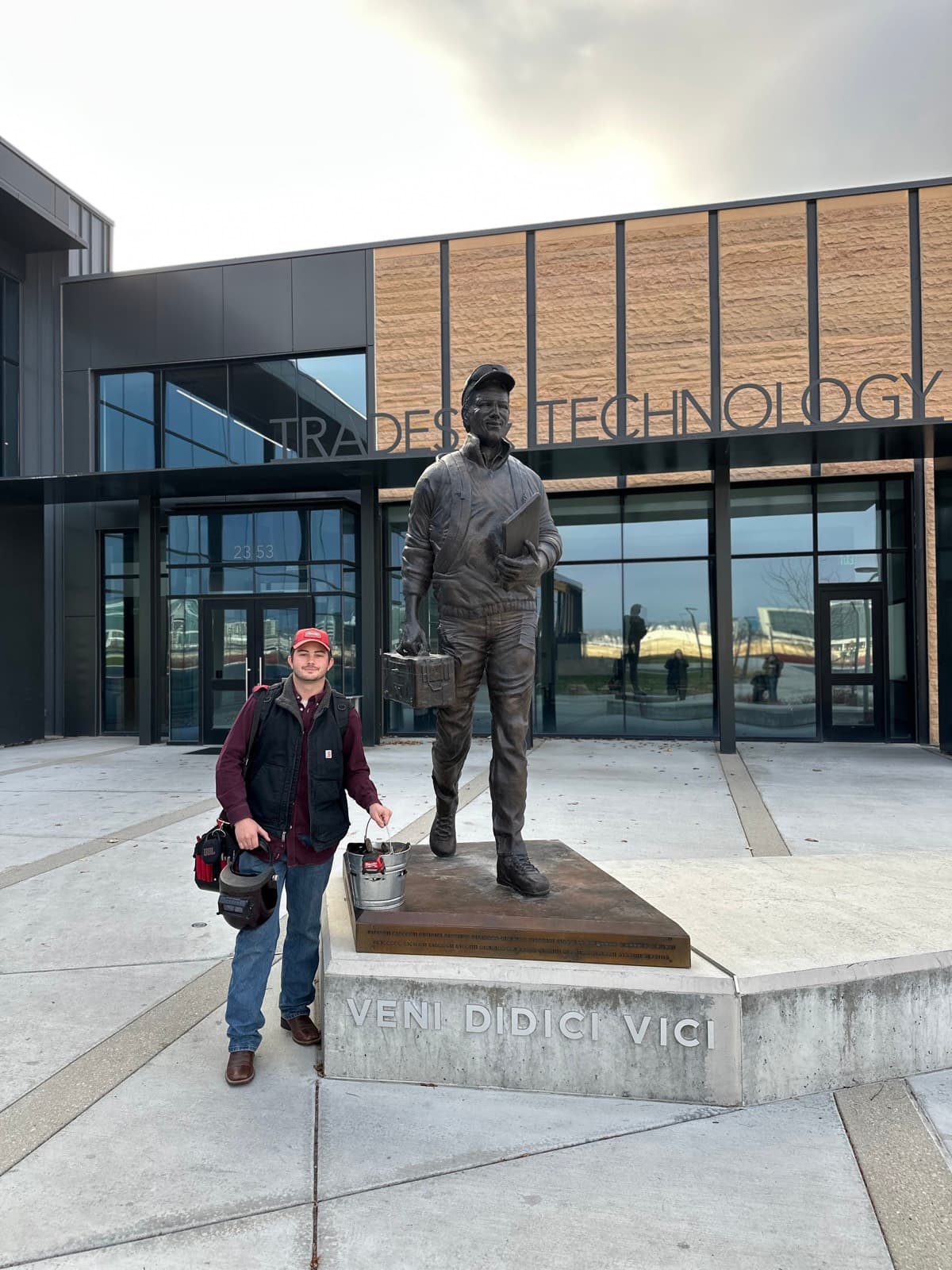 Dylan at M-Tech in front of the Trades and Technology building, beside a bronze tradesman statue